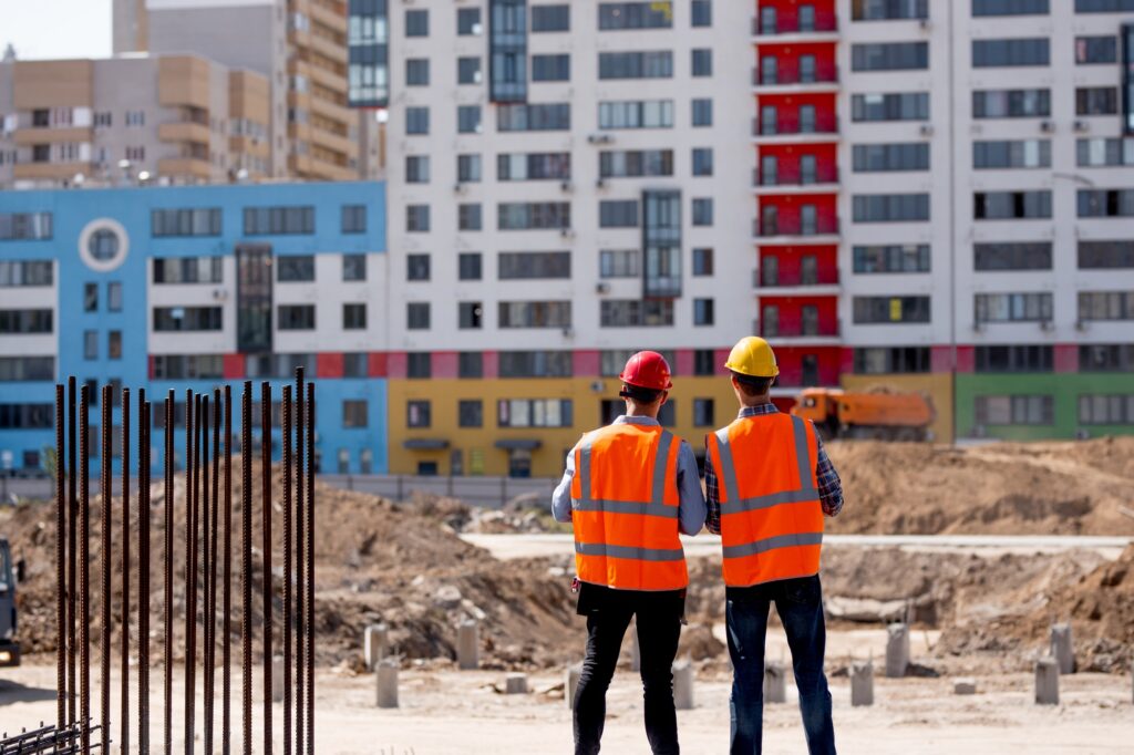 Two men dressed in orange work vests and helmets talk about the construction process on the building